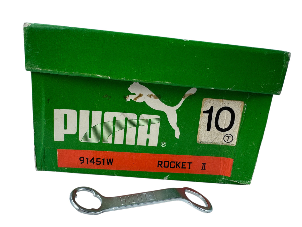 A vintage green Puma Rocket II shoebox with white branding and a red label, shown with a small silver wrench in front on a white background.