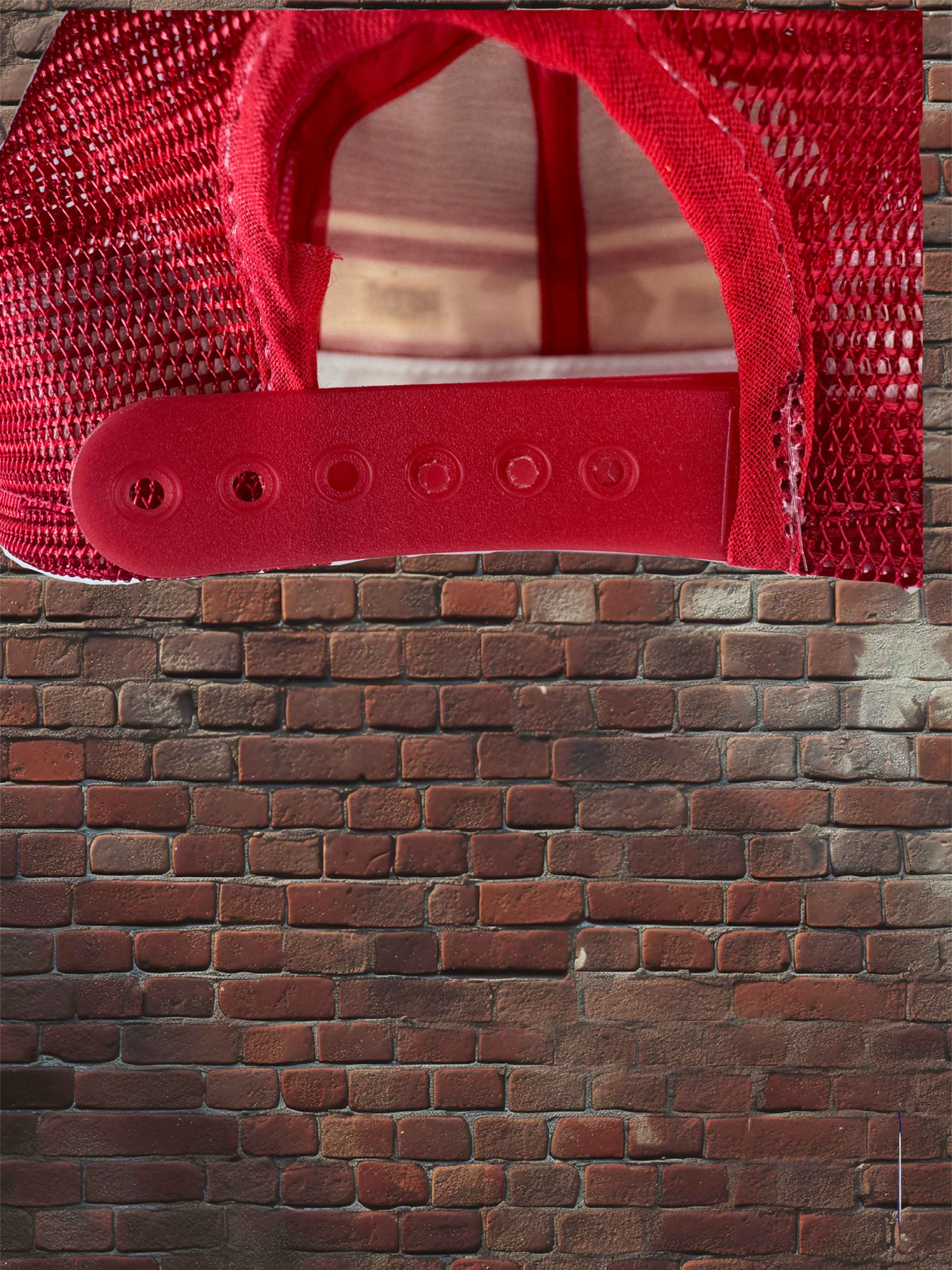 A red trucker-style cap shown from the back, with a red mesh back and red adjustable strap, photographed against a brick wall background.