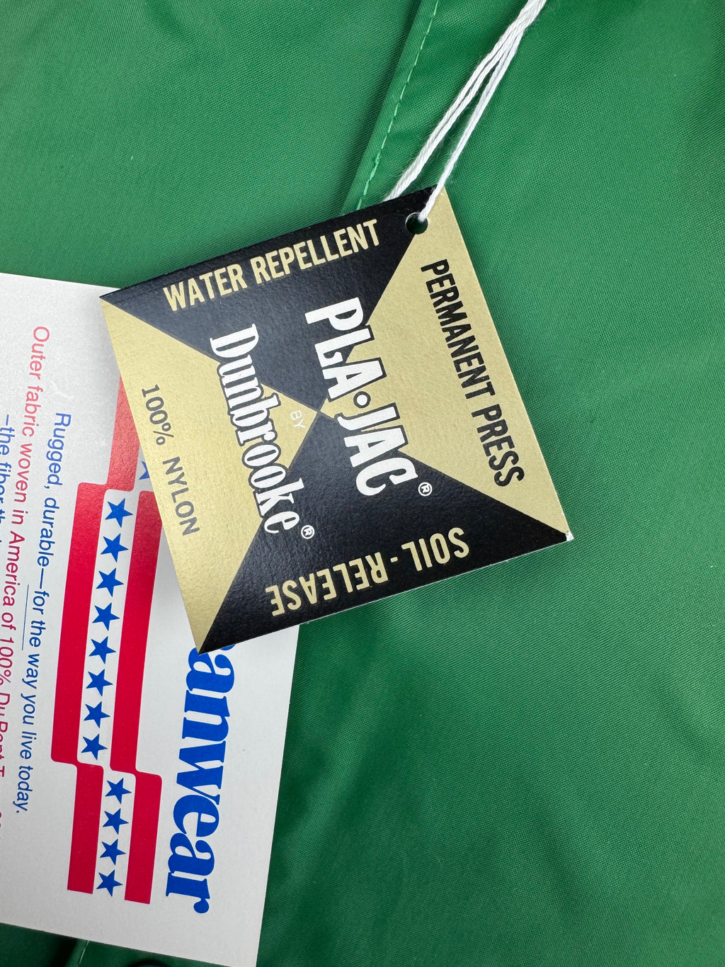 Close-up of a green garment tag with a black and gold hanging label reading 'Dunbrooke Water Repellent' and 'Pla-Jac Permanent Press Soil-Release,' alongside a white Americanwear paper tag.