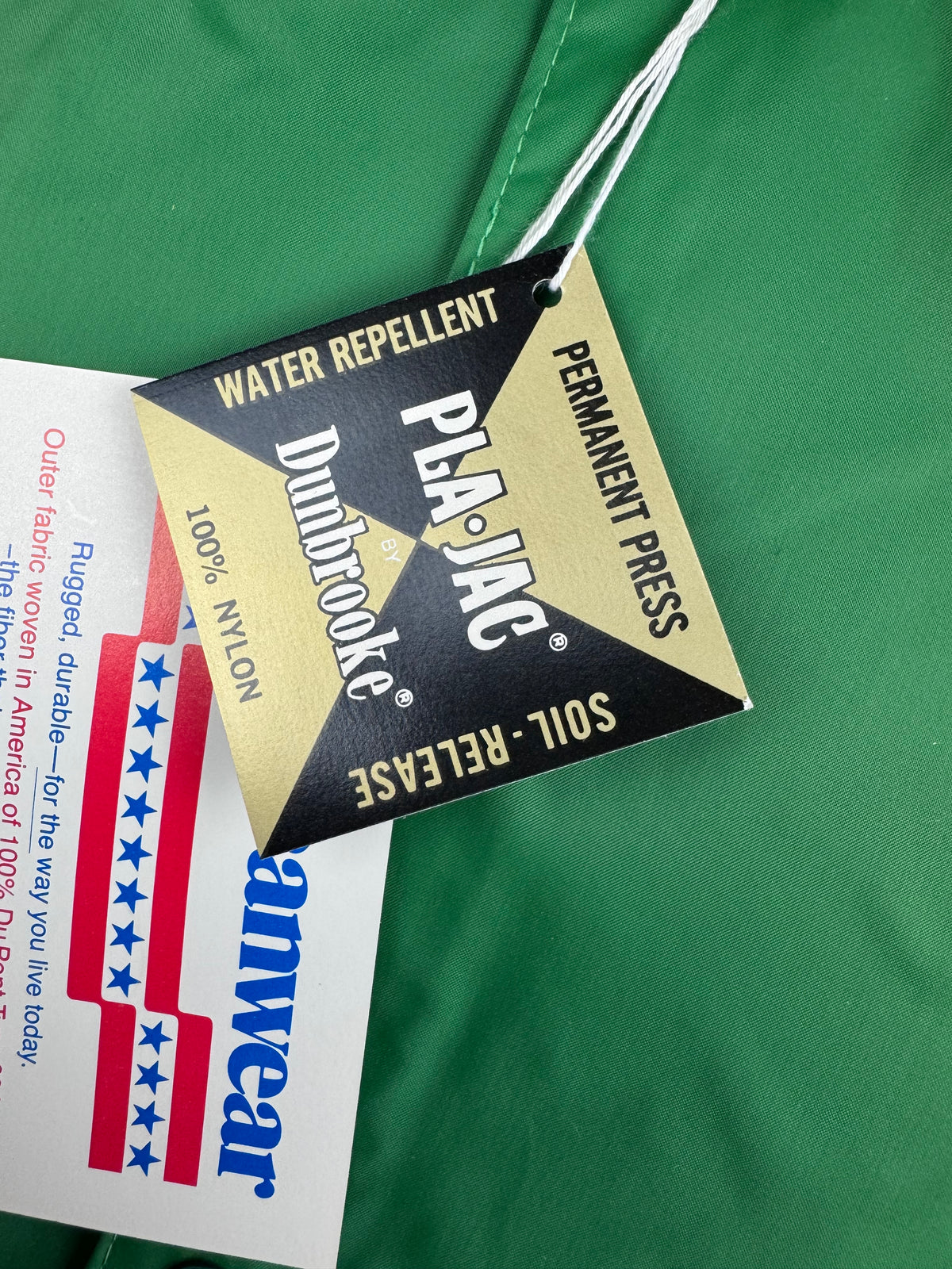 Close-up of a green garment tag with a black and gold hanging label reading 'Dunbrooke Water Repellent' and 'Pla-Jac Permanent Press Soil-Release,' alongside a white Americanwear paper tag.
