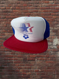 Vintage Los Angeles 1984 Olympics hat with white foam front, Olympic star graphic, Arena logo, red brim, and blue mesh against a brick background.