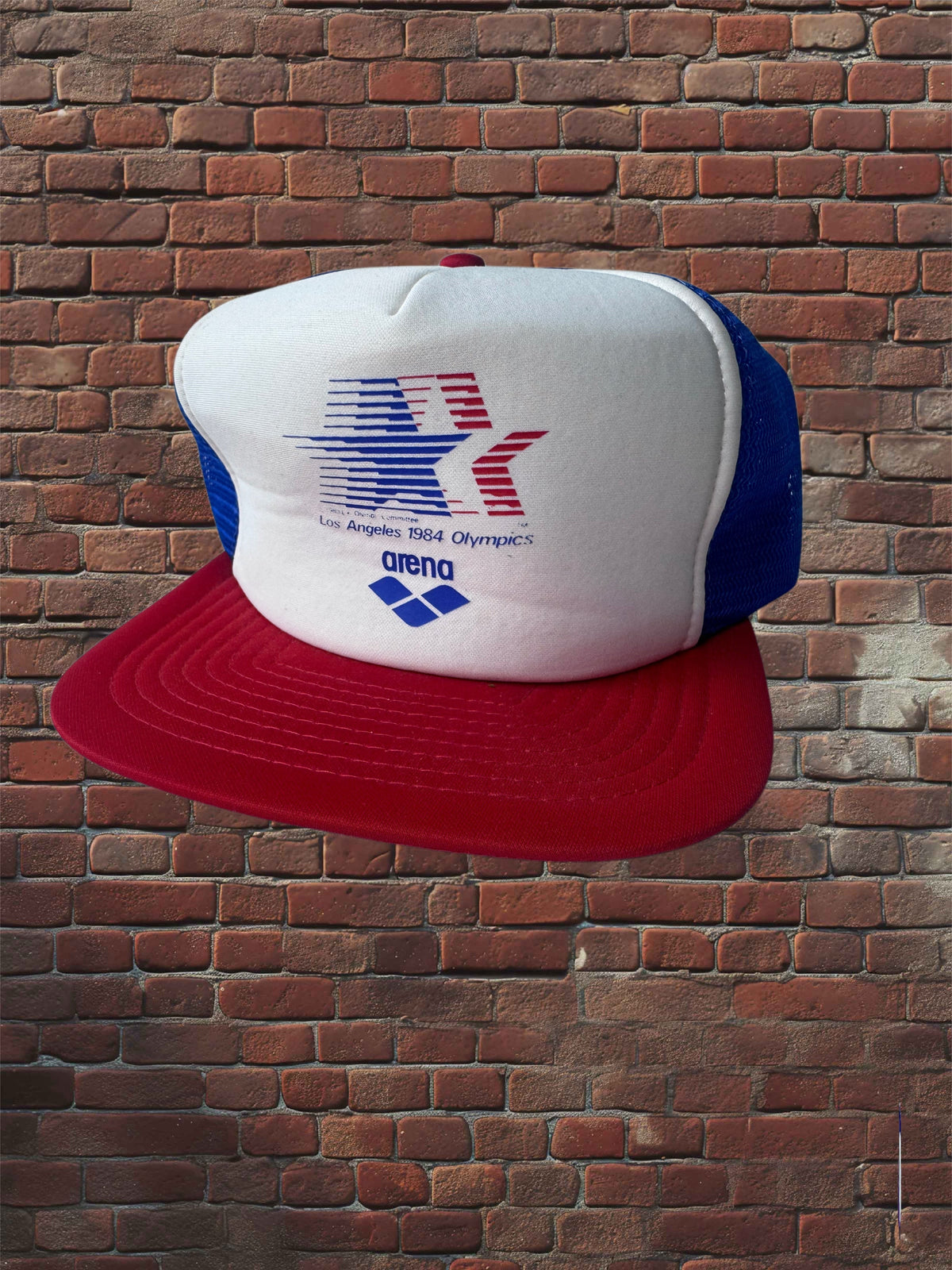 Vintage Los Angeles 1984 Olympics hat with white foam front, Olympic star graphic, Arena logo, red brim, and blue mesh against a brick background.