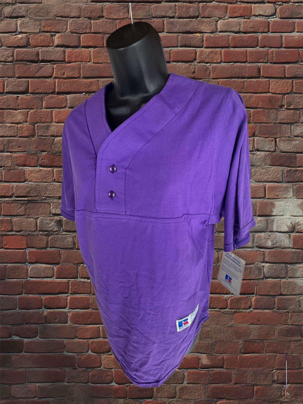 A purple softball shirt with a short button placket and three-quarter sleeves, displayed on a black mannequin torso against a red brick wall background.