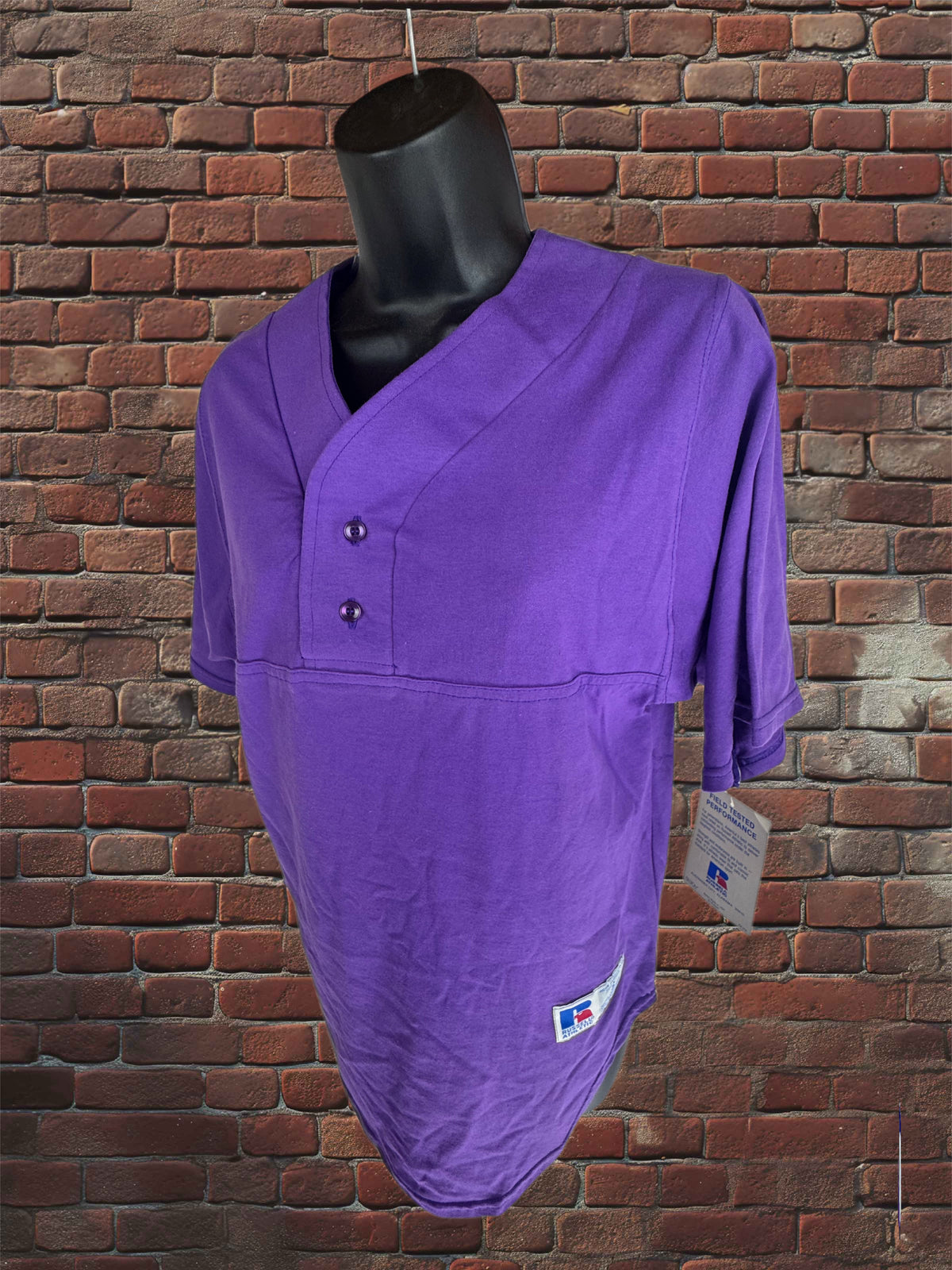 A purple softball shirt with a short button placket and three-quarter sleeves, displayed on a black mannequin torso against a red brick wall background.