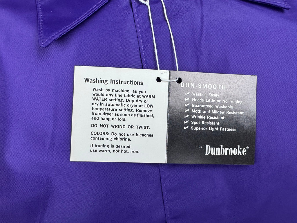A close-up of a purple collared garment with a white hanging tag that reads 'Dunbrooke' and includes washing instructions and care details.