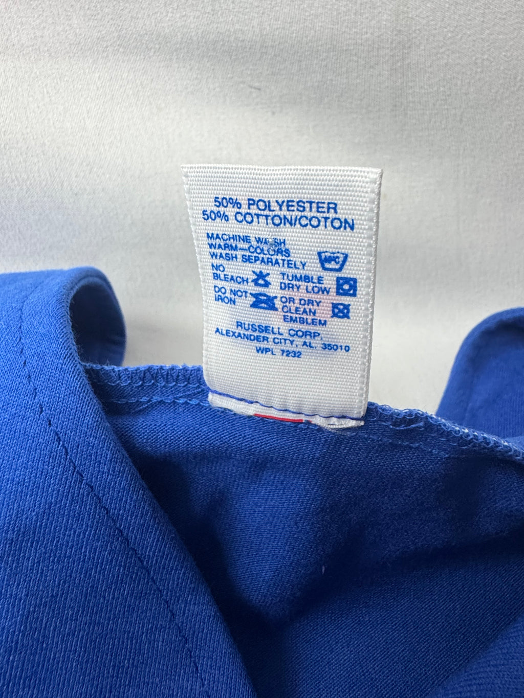 Close-up of a blue garment's inner neck label showing fabric composition as 50% polyester and 50% cotton/cotton, with Russell Corp branding printed on the white tag.