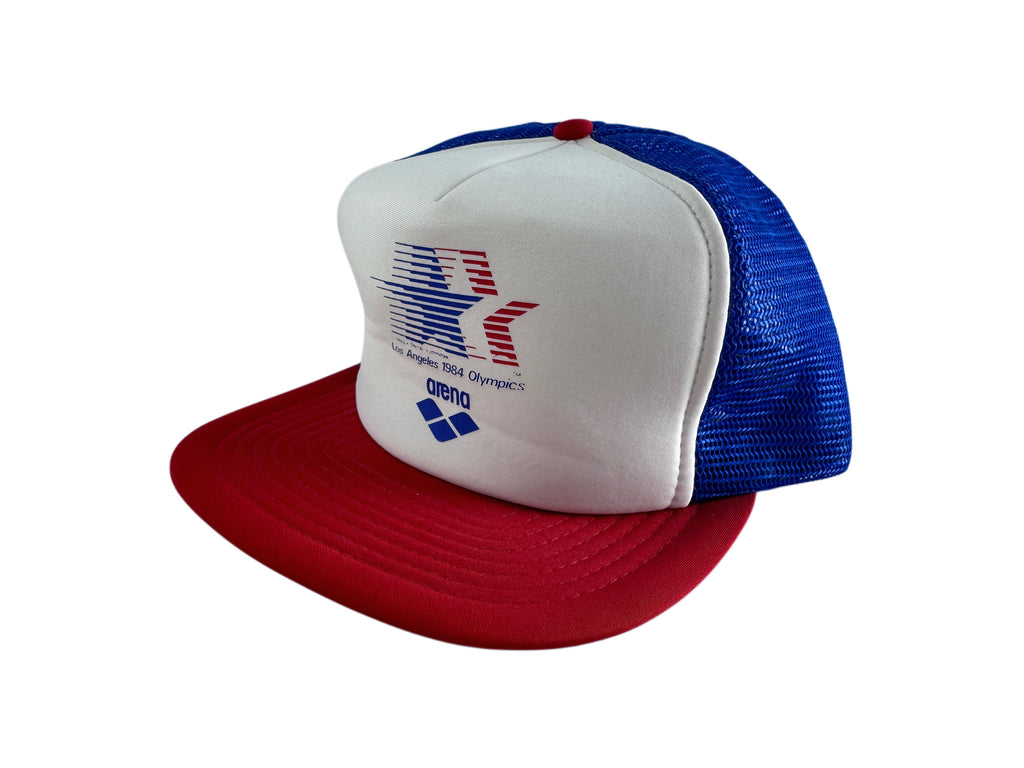 A red, white, and blue trucker hat featuring a white front panel with a red and blue star logo and the text 'Los Angeles 1984 Olympics' and 'arena'. The back of the cap has blue mesh panels, and the flat brim is red.