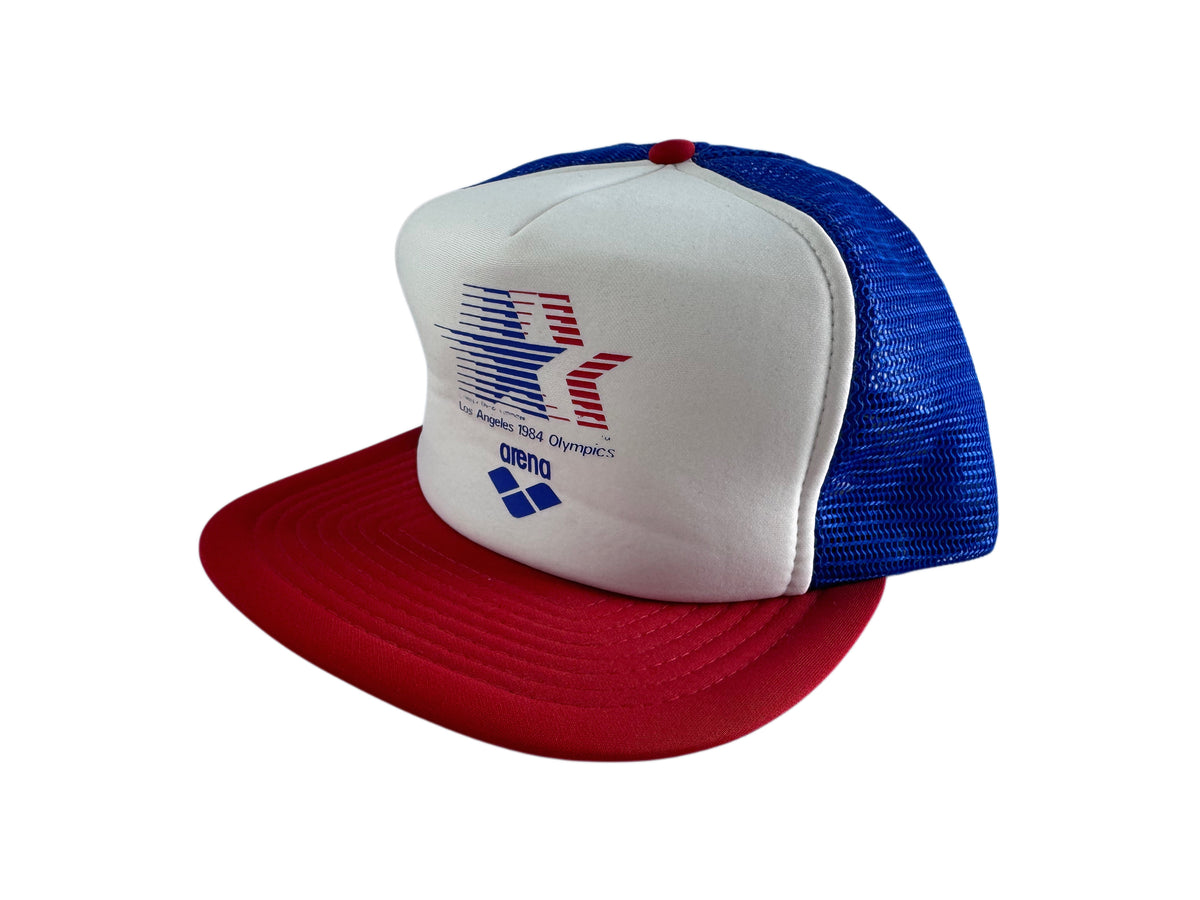 A red, white, and blue trucker hat featuring a white front panel with a red and blue star logo and the text 'Los Angeles 1984 Olympics' and 'arena'. The back of the cap has blue mesh panels, and the flat brim is red.