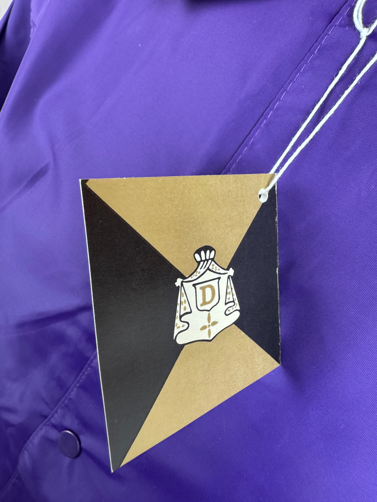 A close-up of a purple garment with a hanging diamond-shaped tag. The tag is black and gold with a white and gold crest featuring the letter 'D'.