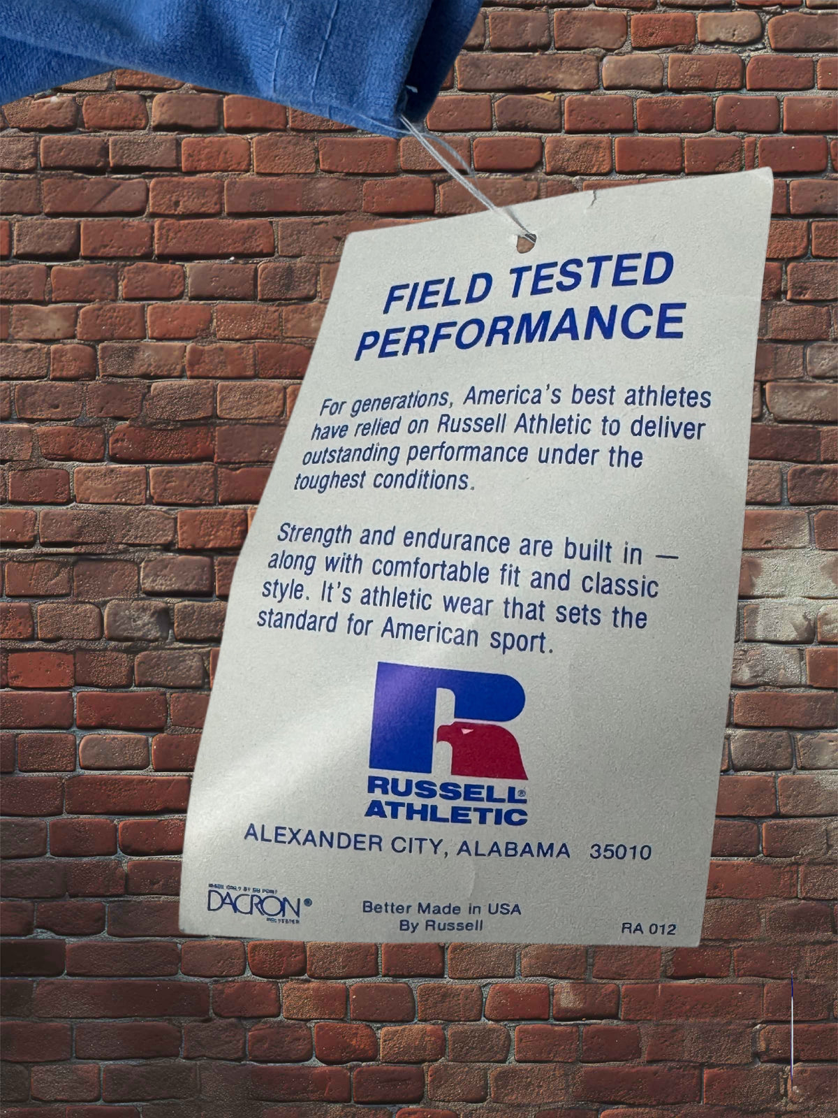 A white paper tag with blue text reading 'FIELD TESTED PERFORMANCE' and the Russell Athletic logo, hanging against a red brick wall.