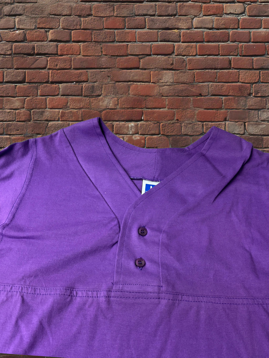 A purple short-sleeve henley shirt with a button placket, laid flat against a red brick wall background.
