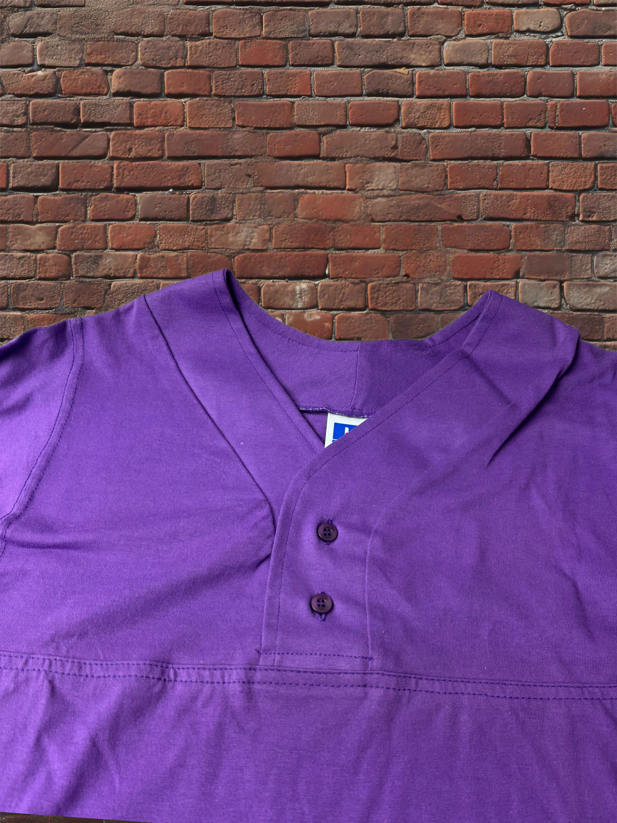 A purple short-sleeve henley shirt with a button placket, laid flat against a red brick wall background.