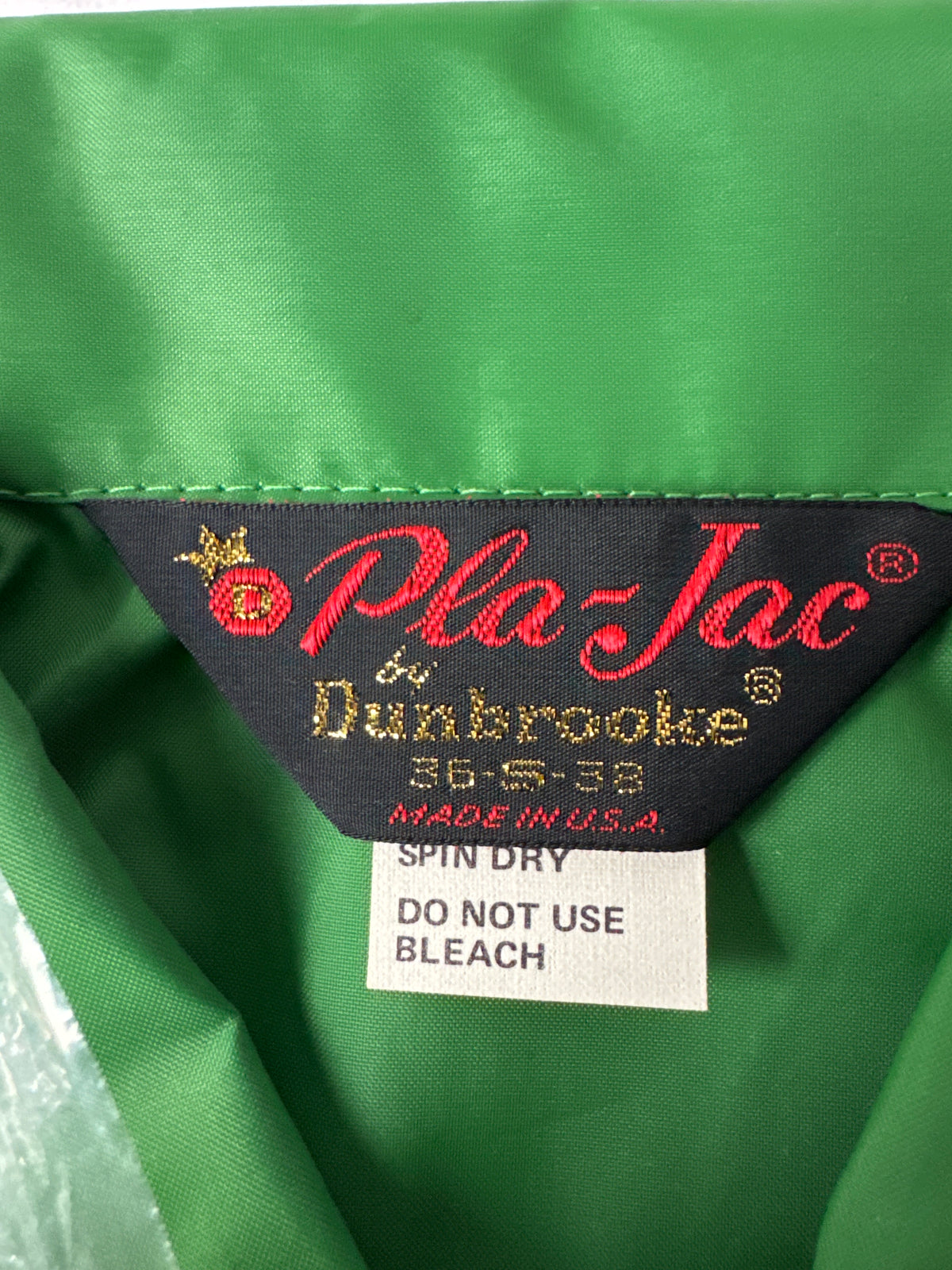 Close-up of a green garment interior showing a black woven label with red and gold text reading 'Pla-Jac by Dunbrooke' and a white care tag below that says 'SPIN DRY DO NOT USE BLEACH'.