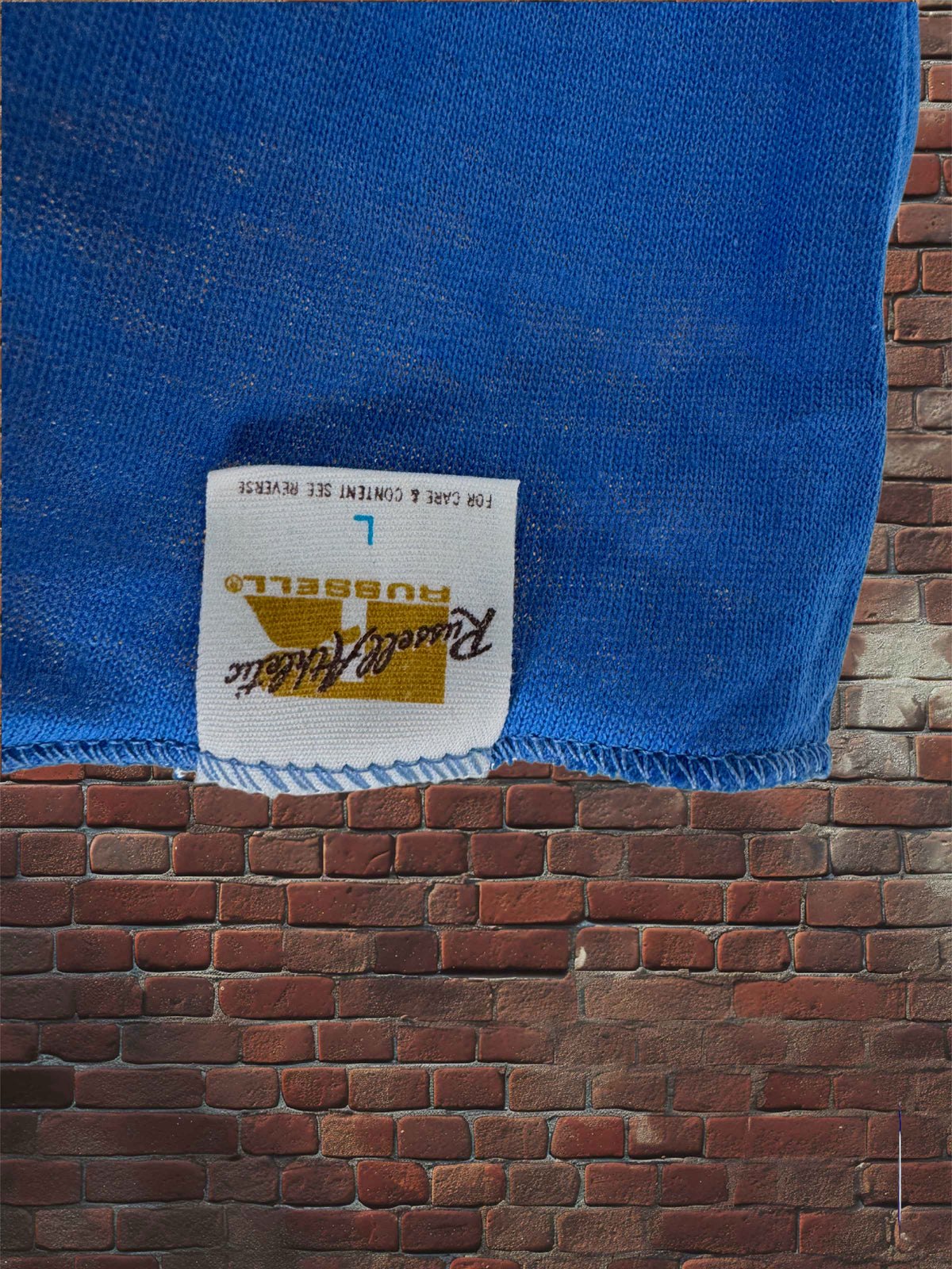 A close-up of a blue knit fabric item with a white sewn-in label reading Russell Athletic, hanging against a red-brown brick wall background.