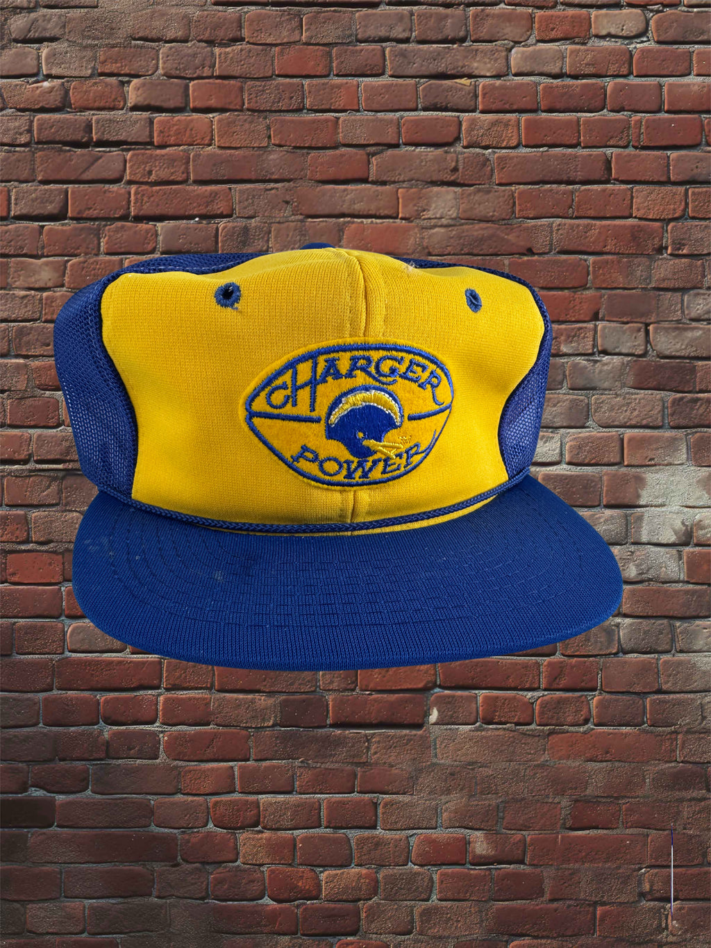 A yellow and blue trucker-style baseball cap with a mesh back and a front patch reading 'Chargers Power,' shown against a red-brown brick wall background.
